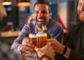 Happy mid adult friends clinking with beer mugs in pub. Three cheerful guys drinking draft beer, celebrating meeting and smiling. Laughing young men enjoying cold pint of beer during night at bar.