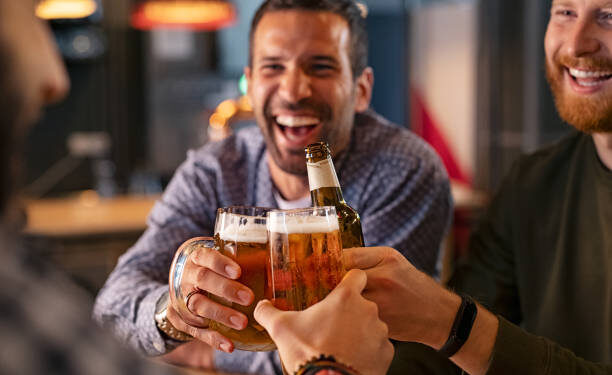 Happy mid adult friends clinking with beer mugs in pub. Three cheerful guys drinking draft beer, celebrating meeting and smiling. Laughing young men enjoying cold pint of beer during night at bar.