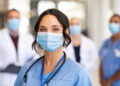 Close up face of confident female nurse in front of his medical staff looking at camera while wearing protective face mask due to covid-19 virus. Smiling surgeon standing  with team in background.