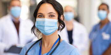 Close up face of confident female nurse in front of his medical staff looking at camera while wearing protective face mask due to covid-19 virus. Smiling surgeon standing  with team in background.