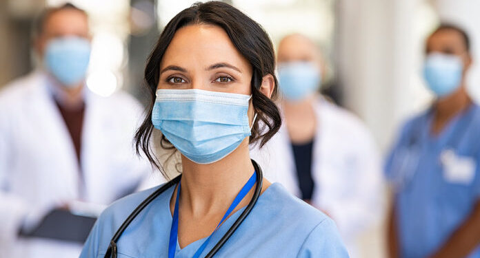 Close up face of confident female nurse in front of his medical staff looking at camera while wearing protective face mask due to covid-19 virus. Smiling surgeon standing  with team in background.