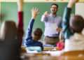 Happy male teacher aiming at his students who raised their hands to answer the question on a class at elementary school.