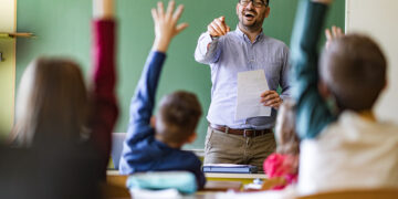 Happy male teacher aiming at his students who raised their hands to answer the question on a class at elementary school.