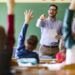 Happy male teacher aiming at his students who raised their hands to answer the question on a class at elementary school.